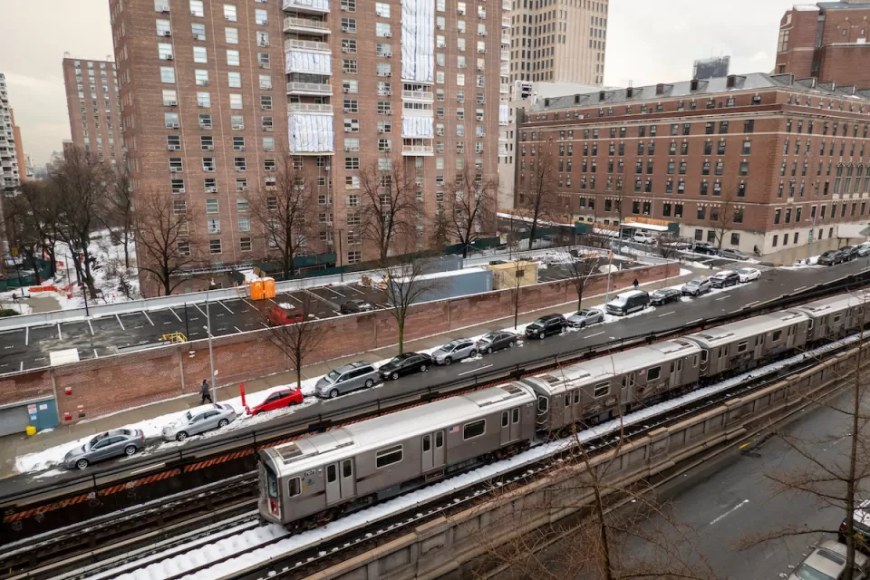 Silver New York City subway train on above ground track with red brick multistory buildings in background.